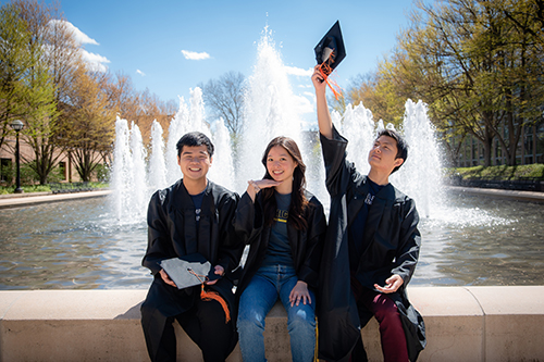 3 students in grad cap and gowns sit in front of the Lurie fountain on north campus in spring